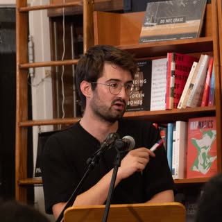 Man on the podium with a microphone and a bookshelf in the background