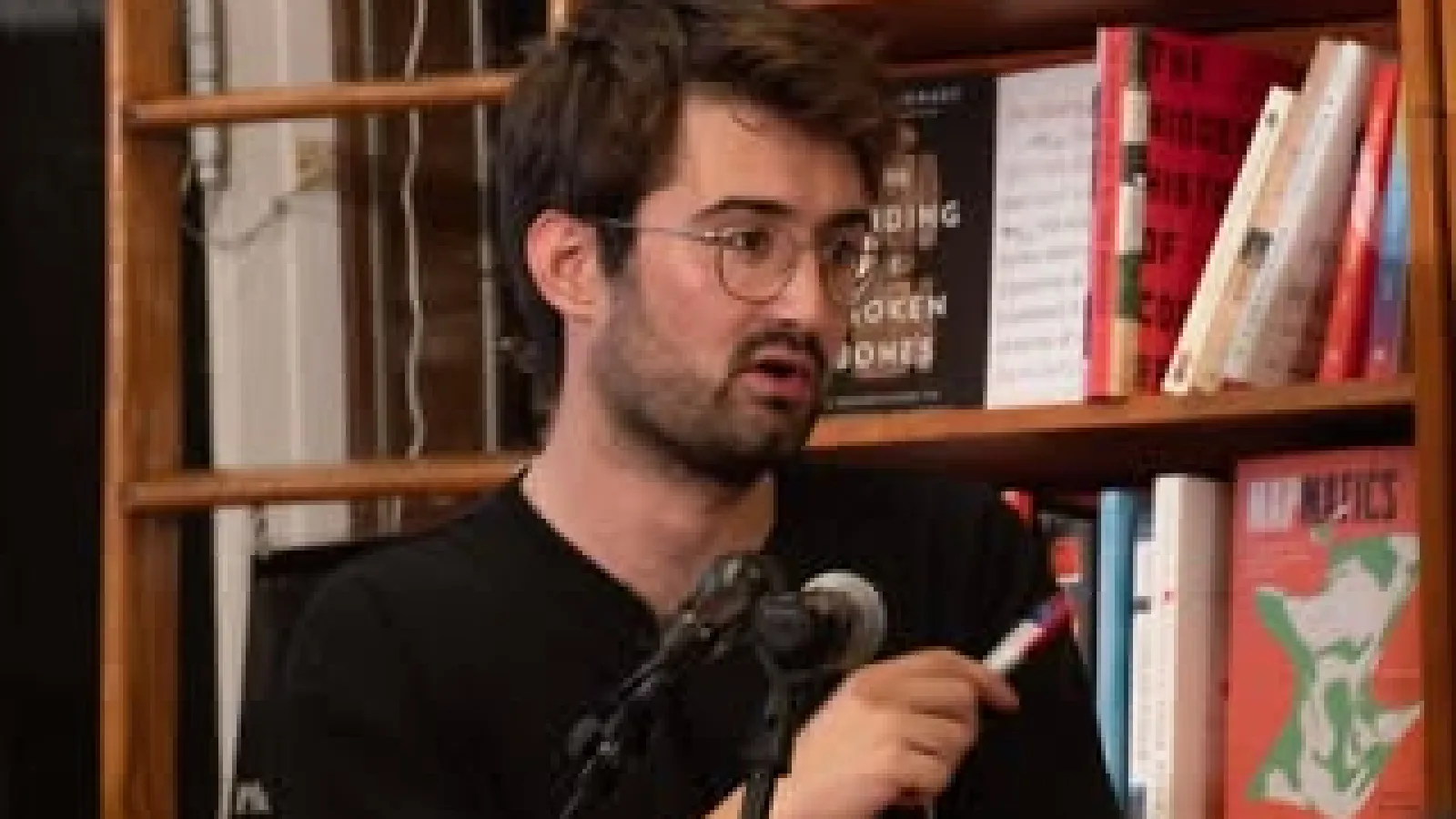 Man on the podium with a microphone and a bookshelf in the background