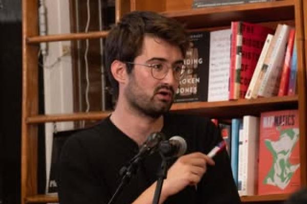 Man on the podium with a microphone and a bookshelf in the background